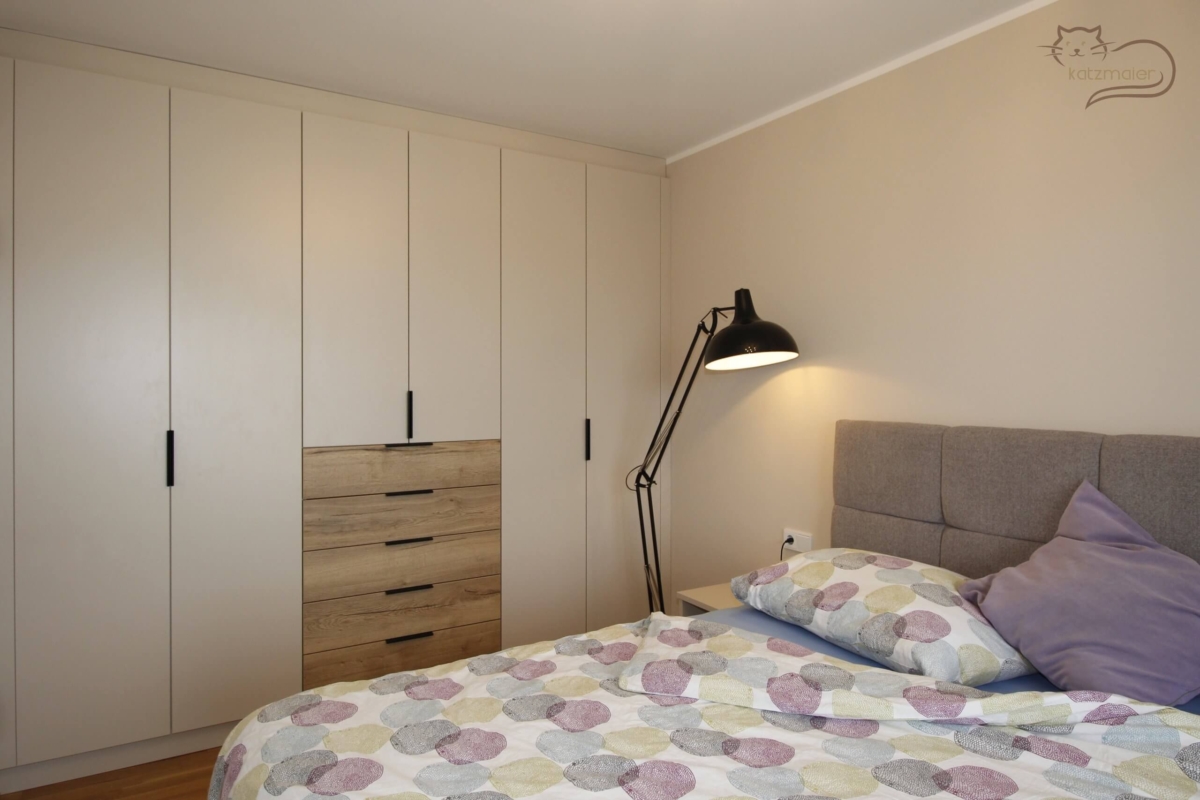 Ein Schlafzimmer mit einem grauen Boxspringbett im Vordergrund. Links vom Bett ein Nachtkästchen in einem warmen Hellgrau und eine große schwarze Stehleuchte. Dahinter an der Wand ist ein raumhoher Schrankverbau mit rechts und links ein Schrank mit je zwei Türen in einem warmen Hellgrau. In der Mitte vom Schrankverbau ein Schrank mit unten fünf breit Laden in Holzoptik und darüber zwei Türen in einem warmen Hellgrau. Die Schränke sind mit schwarzen Griffen bedienbar.