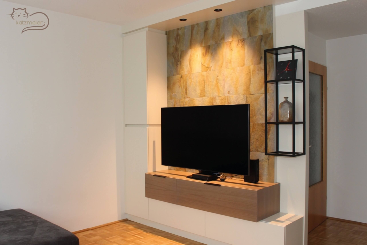 TV-Wandverbau in Weiß kombiniert mit Möbelteilen in Eiche furniert. Links an der Mauerseite ein raumhoher Schrank in Weiß. Der Schrank hat zwei Türen, die mit einer Griffmulde bedienbar sind. Rechts vom Schrank ein Unterschrank in Weiß mit zwei Laden. Die beiden Laden sind grifflos mit Tip-On bedienbar. Darüber ein aufgesetzter Unterschrank in Eiche mit zwei Klappen. Die beiden Klappen sind mit schwarzen Griffleisten bedienbar. Hinter dem TV-Verbau eine Wandverkleidung mit einer braunen und grauen Steinfurnier. Rechts von der Wandverkleidung hängt ein offenes Metallregal in Schwarz.