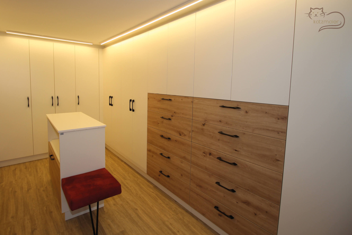 Ein begehbarer Kleiderschrank in Weiß kombiniert mit Möbelteilen in Eiche. Der Kleiderschrank besteht aus Schränken mit Türen in Weiß und aus Schränken mit unten Laden in Eiche und darüber Türen. Die Schränke sind mit schwarzen Griffen bedienbar. In der Mitte vom Schrankraum ist eine halbhohe Ablage in Weiß mit einer kleinen Sitzmöglichkeit. Die Sitzpolsterung ist mit einem roten Stoff tapeziert. An der Decke entlang von den Schränken sind LED-Lichtleisten.