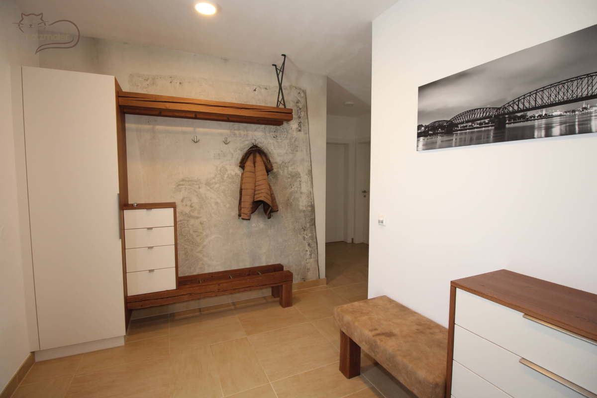 Ein Vorzimmer mit Möbeln in weißen Dekorplatten und bearbeiteten Altholzbalken in Fichte. Links ein geschlossener Kleiderschrank in Weiß. Daneben eine Sitzfläche aus zwei Altholzbalken. Auf den Altholzbalken ein Ladenkastl in Weiß. Oben zwei Altholzbalken als Hutbord. Dahinter an der Wand eine moderne Wandtapete.