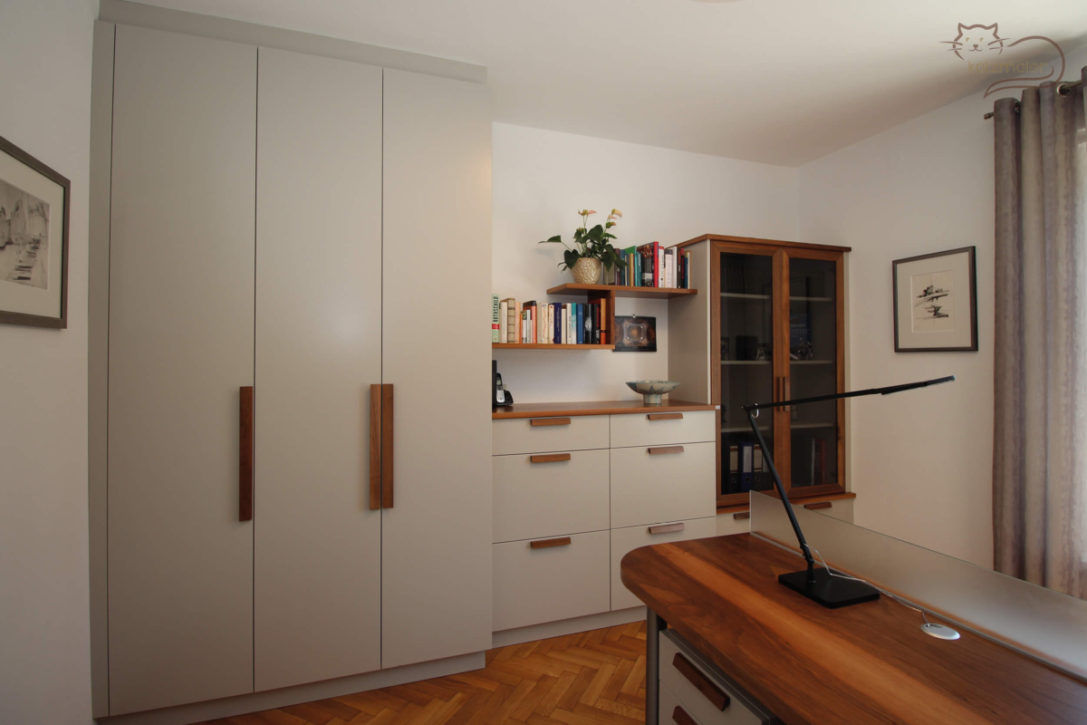Ein Arbeitszimmer mit einer Erinrichtung in Nuss furniert kombiniet mit Möbelteilen in einem hellen warmen Grau. Links von der Mauer ein raumhoher 3-türiger Schrank in Grau.Mittig ein halbhoher Ladenschrank in Creme mit einer Einfassung in Nuss furniert. Darüber ein versetztes Bücherregal in Nuss furniert. Rechts daneben ein Unterschrank mit einem aufgeseten Schrank in Creme. Der Schrank hat zwei Rahmentüren in Nuss mit einer Glasfüllung.