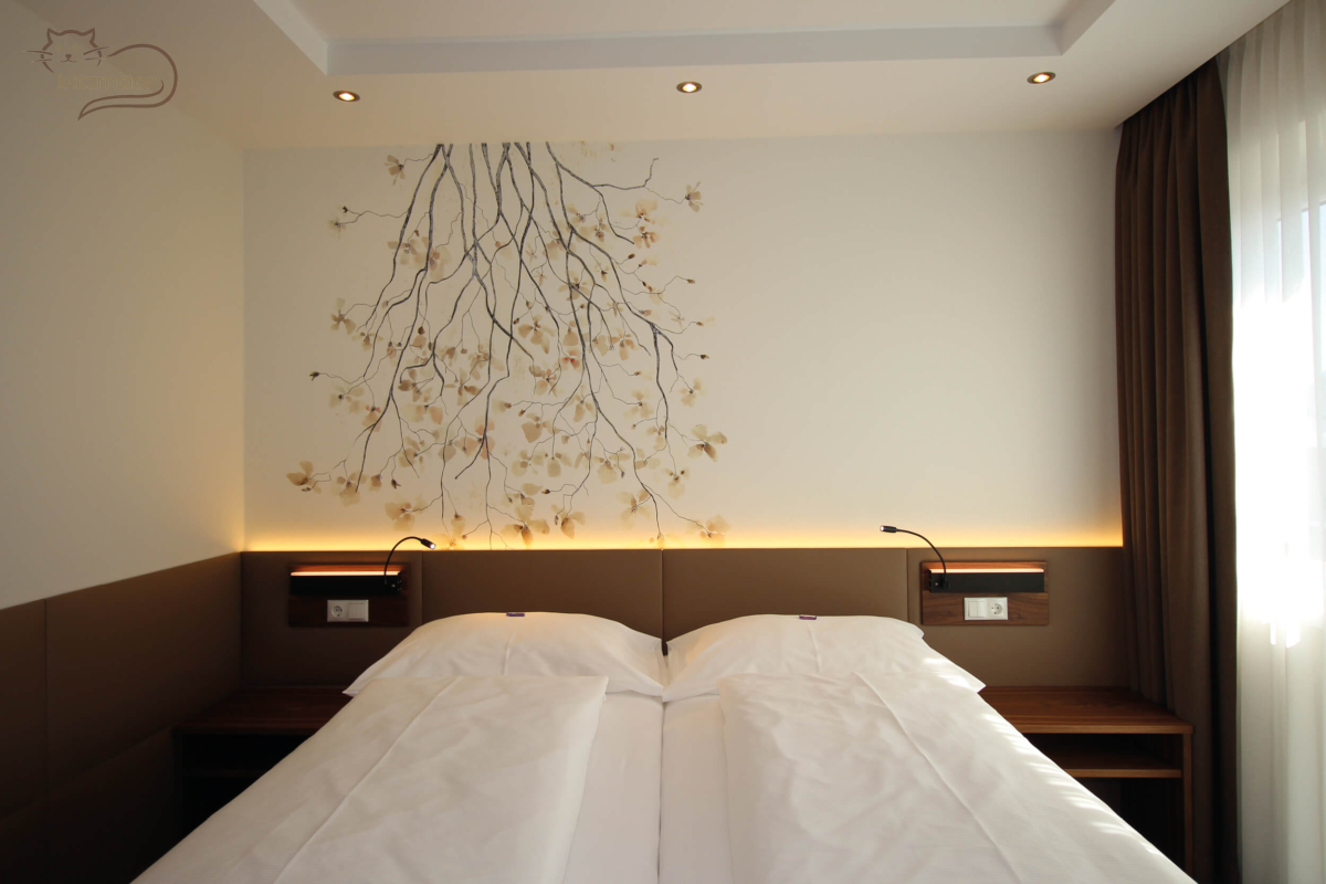 Ein Gästezimmer mit einem großen Doppelbett. Rechts und links davon ein Nachtkästchen in Nuss furniert. Hinter dem Bett und auf der linken Seite eine Wandverkleidung mit tapezierten Lederfeldern in Braun. Auf der Wandverkleidung über den Nachtkästchen eine schwarze Wandleuchte auf eine furnierten Nussplatte. Hinter der Wandverkleidung eine eingefräste LED-Beleuchtung. Auf der gesamten Wand dahinter eine weiße Wandtapet mit einem großen Blumenstrauch mit gelben Blumenblüten auf der linken Seite.