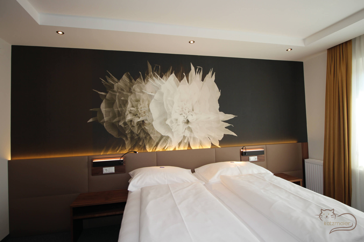 Ein Gästezimmer mit einem großen Doppelbett. Rechts und links davon ein Nachtkästchen in Nuss furniert. Hinter dem Bett eine Wandverkleidung mit tapezierten Lederfeldern in Braun. Auf der Wandverkleidung über den Nachtkästchen eine schwarze Wandleuchte auf eine furnierten Nussplatte. Hinter der Wandverkleidung eine eingefräste LED-Beleuchtung. Auf der gesamten Wand dahinter eine schwarze Wandtapet mit mittig eine große weiße Blumenblüte.