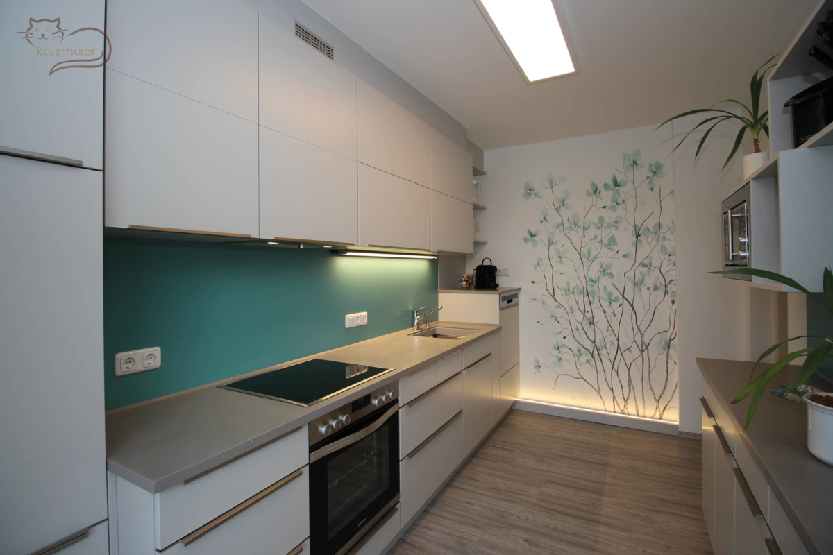 Eine Küchenzeile in Birkengrau kombiniert mit einer Rückwand in Petrol. Die Küchenzeile besteht aus Unterschränken und Oberschränken mit Klappfronten. Die Küchenfronten sind mit Griffleisten in Edelstahl bedienbar. Am Ende der Küchenzeile befindet sich eine Wandtapete mit petrolfarbenen Blütenblättern. Die Wandtapete wird von unten beleuchtet.