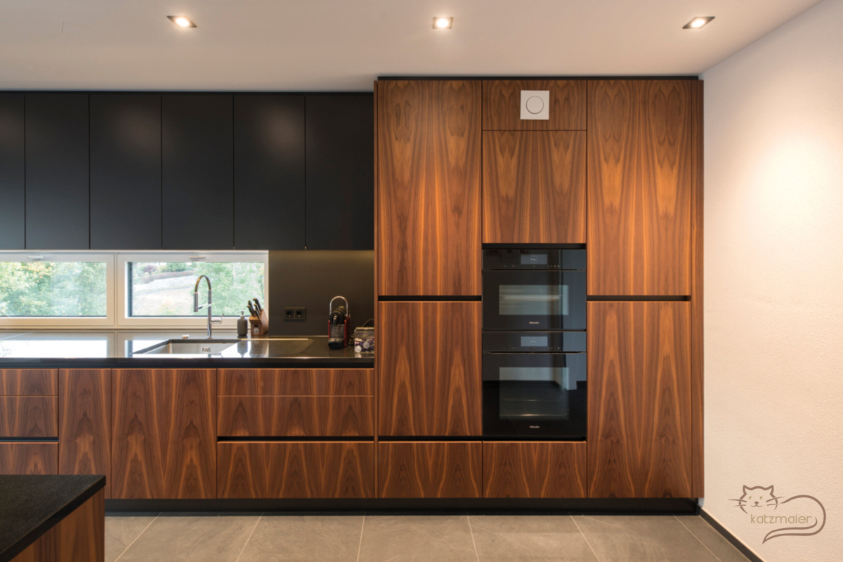 Schwarze Küchenzeile kombiniert mit Küchenfronten in Nussfurnier stehend. Die Küchenzeile besteht aus Unterschränken mit darüber Oberschränken und Hochschränken.