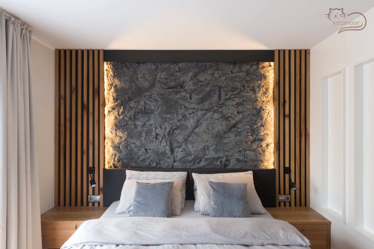 Schlafzimmer mit Möbeln in Eiche. In der Mitte steht ein Bett in Eiche mit einem tapezierten Kopfhaupt in Dunkelgrau. Links und rechts vom Bett ist ein Nachtkästchen mit einer Lade. Hinter dem Bett ist ein Wandverbau. Links und rechts an der Mauer befinden sich Eichensprossen. In den Eichensprossen sind auf beiden Seiten schwarze Leuchten und Steckdosen mit Lichtschalter verbaut. An der Mauer zwischen den Eichensprossen befindet sich eine Wandverkleidung in einer grauen Felsoptik. Die Felswand wird links und rechts von den Eichensprossen indirekt beleuchtet.