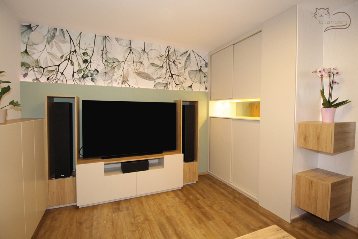 Eine Wohnzimmereinrichtung in einem warmen Hellgrau und Eiche. Auf der linken Seite ist ein Highbordkastl in Hellgrau. Anschließend befindet sich der Fernsehverbau in Hellgrau und Eiche. Links und rechts vom Fernsehunterschrank sind offene Regale in Eiche. Die Wandfarbe hinter dem Fernseher ist in einem Pastellgrün und im oberen Bereich ist eine Wandtapete mit großen Blättern und Blüten in verschiedenen Grüntönen. Auf der rechten Seite befindet sich ein raumhoher Wandverbau in Hellgrau. Der Wandverbau besteht aus einem Schrank mit Schiebetüren. In der Mitte ist eine offene Nische mit einem Spiegel und einer indirekten Beleuchtung. Am Ende vom Wandverbau sind zwei Hängekastl in Eiche.