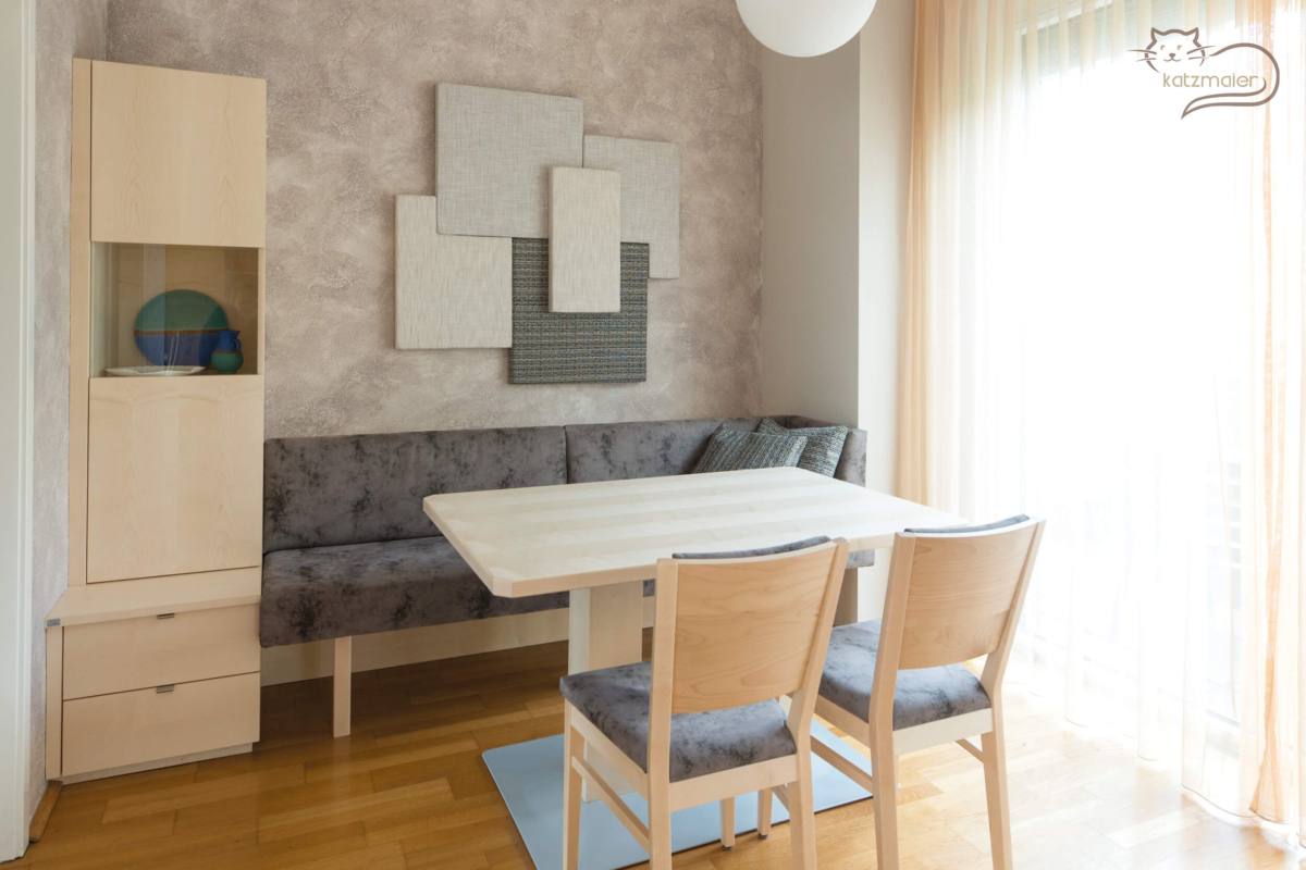 Ein Essbereich in Ahorn furniert mit einer großen Fensterfront auf der rechten Seite. Auf der linken Seite befindet sich direkt an der Wand ein Unterschrank mit zwei Laden und darauf eine seichterer Aufsatzschrank. Anschließend ist eine Essbank mit einem grauen Stoff tapeziert. Davor steht der Esstisch mit einer Esstischplatte in Ahorn. Gegenüber der Bank stehen zwei Sessel in Ahorn und mit einem grauen Stoff tapeziert. Die Wand hinter dem Schrank und der Essbank ist in einem warmen Grauton gestrichen. Über der Essbank hängt ein Akustikbild, das mit verschiedenen Stoffen tapeziert ist.