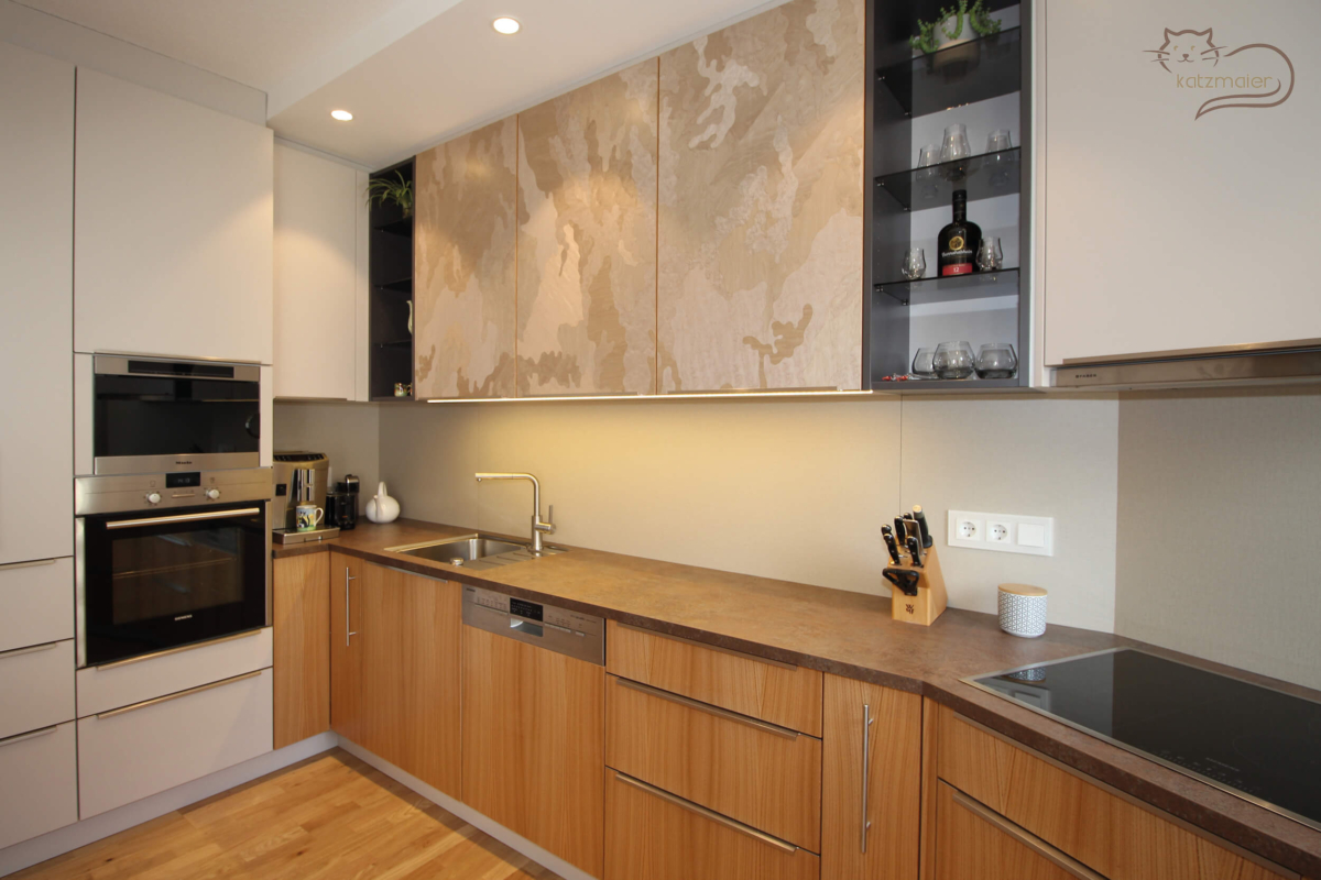 Eine Küchenzeile in hellen Dekorplatten kombiniert mit Fronten in Kastanie furniert. Die Fronten der Oberteile sind mit einem grauen Kunstfurnier in Camoflage-Optik furniert.