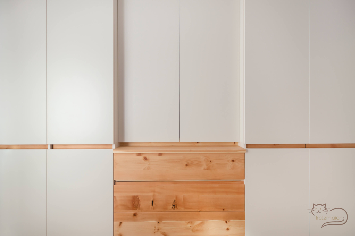 Wandansicht eines Kleiderschrankes in Creme und Zirbe massiv. In der Mitte unten Laden mit Zirbenfronten. Darüber ein seichterer Aufsatzschrank in Creme. Rechts und links davon ein Kleiderschrank in Creme. Die Schränke sind mit einer Griffleiste in Zirbe bedienbar.