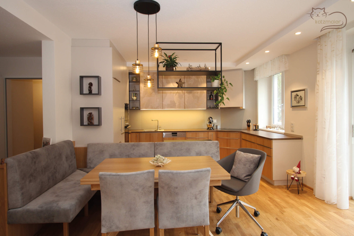 Im Vordergrund eine Essgruppe mit einer Eckbank und 3 Stühlen. Die Eckbank und Stühle sind in einem hellen Grauton tapeziert. Über dem Esstisch hängt eine schwarze Hängeleuchte. Hinter der Essgruppe befindet sich eine Küche in hellem Dekorplatten kombiniert mit Fronten in Kastanie furniert.