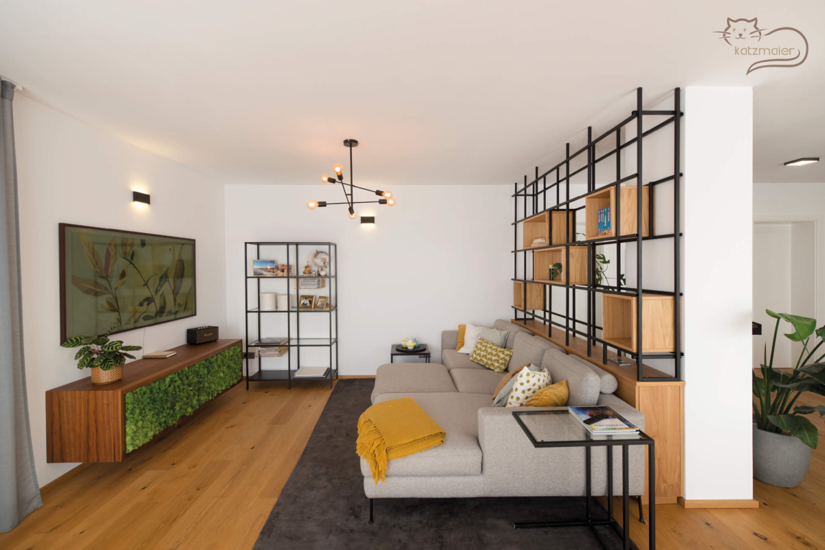 Ein großzügiger helles Wohnzimmer. Auf der rechten Seite an der Wand ein aufgehängter TV-Unterschrank mit einer Einfassung in Nuss. 2 Rahmentüren mit einem künstlich bearbeiteten Moos als Füllung. Links vom Foto ein schwarzes Metallregal mit offenen Nischen in Eiche als Raumteiler. Davor steht eine graue Couch mit Zierkissen.