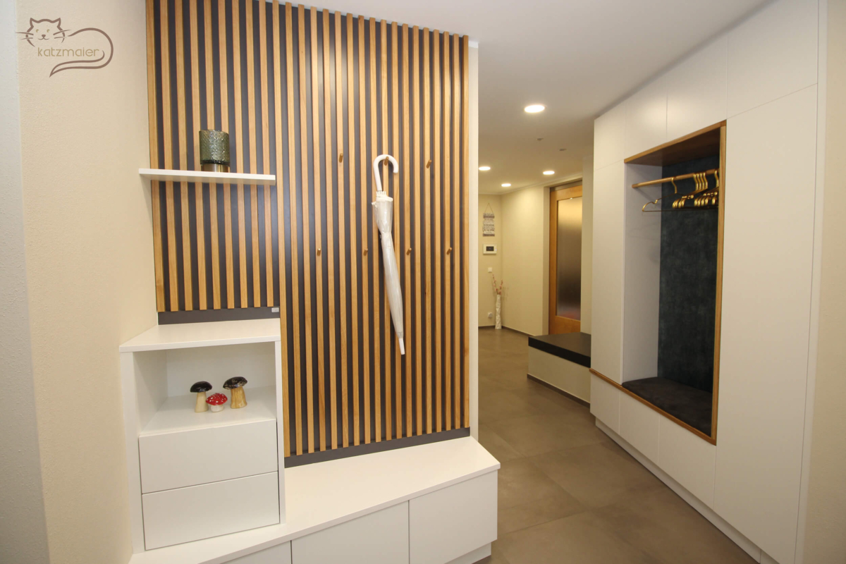 Ein Vorzimmer mit Möbel in Weiß kombiniert mit Möbelteile in Eiche. Auf der linken Seite eine Vorzimmergarderobe mit einem abgeschrägten Unterschrank in Weiß. Links an der Mauerseite steht auf dem Unterschrank ein Aufsatzschrank mit unten zwei Laden und oben einer offenen Nische in Weiß. Dahinter eine Wandverkleidung mit Eichensprossen und einem Ablagebord in Weiß. Die Wandverkleidung mit den Eichensprossen dient als Garderobe. Dahinter auf der rechten Mauerseite befindet sich ein abgeschrägter raumhoher Schrank mit mittig einer offenen Nische. Die Einfassung der Nische ist in Eiche. In der Nische befindet sich ein samtbraun tapezierter Sitzpolster. Die Rückwand der Nische ist mit einem samtblauen Stoff tapeziert. In der Nische ist eine Holzkleiderstange befestigt mit goldenen Kleiderbügeln.