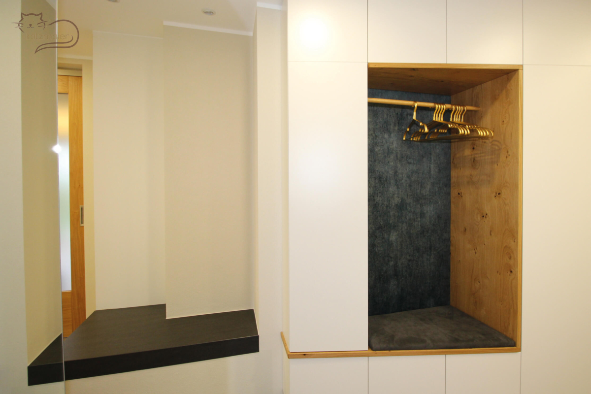 Auf der linken Seite eine Vorzimmergarderobe mit einem abgeschrägten Schrank in Weiß mit mittig einer offenen Nische. Die Einfassung der Nische ist in Eiche. In der Nische befindet sich ein samtbraun tapezierter Sitzpolster. Die Rückwand der Nische ist mit einem samtblauen Stoff tapeziert. In der Nische ist eine Holzkleiderstange befestigt mit goldenen Kleiderbügeln.