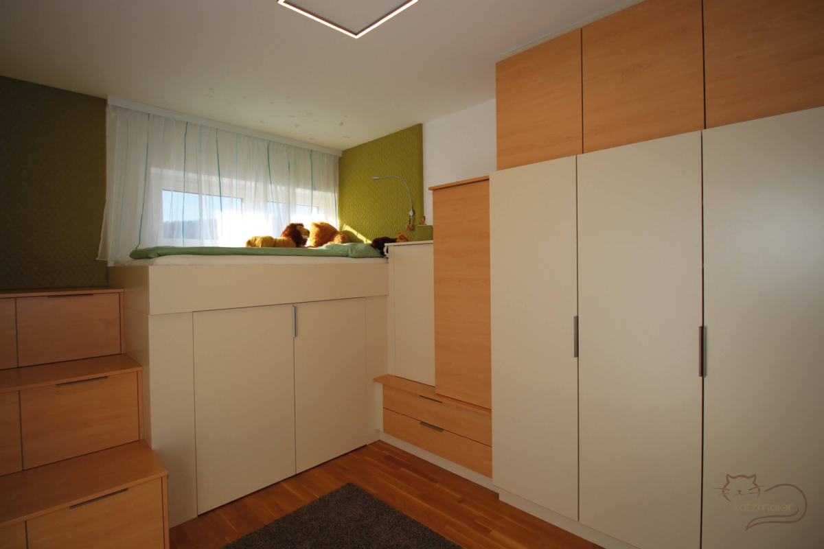 Ein Kinderzimmer in Hellbeige und Buche beschichtet. Auf der linken Mauerseite befindet sich der Stiegenaufgang in Buche zum Hochbett. Die Stiegen werden gleichzeitig auch als Laden verwendet. Der Hochbettverbau ist in Hellbeige. Das Kopfhaupt vom Bett ist eine tapezierte Wandverkleidung in Grün. Unter dem Hochbett sind zwei Türen zum Öffnen und dahinter ist genügend Platz für Stauraum. Auf der rechten Seite entlang der Mauer befindet sich ein Schrankverbau in Hellbeige und Buche kombiniert.