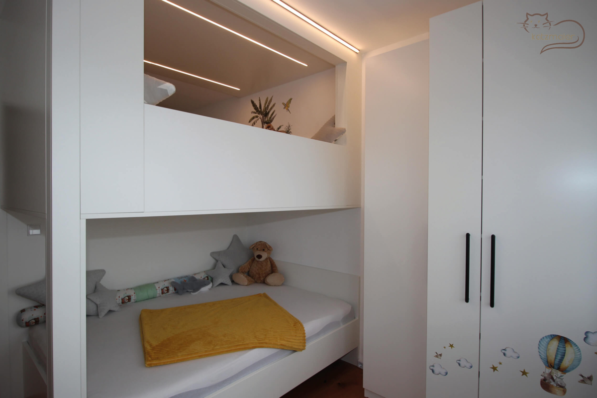 Kinderzimmer in weißen Dekorplatten. Auf der linken Seite ein Hochbett. Rechts davon zwei tiefenversetzte Kleiderschränke in Weiß, die mit schwarzen Griffen bedienbar sind.