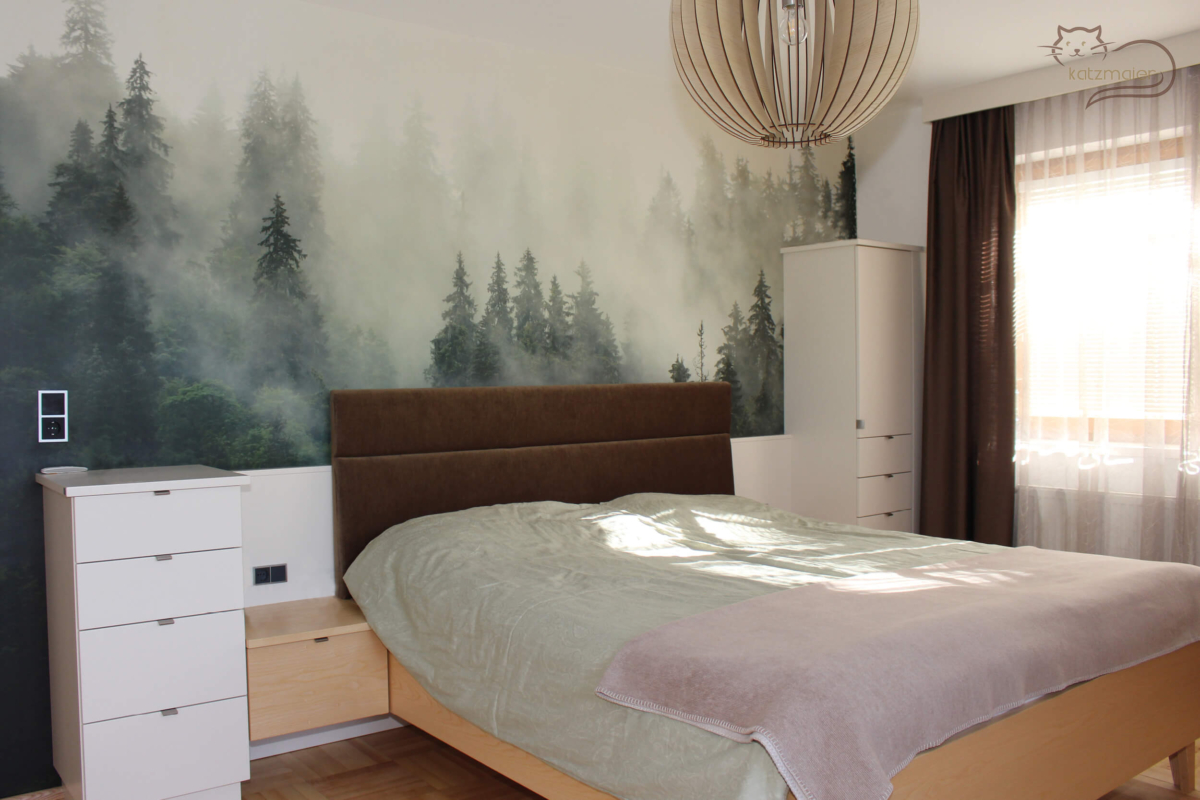 Ein Schlafzimmer in Esche furniert kombiniert mit Möbelteilen in Weiß. Das Bett und die aufgehängten Nachtkästchen sind in Esche. Hinter dem Bett ist eine weiße Wandverkleidung und ein tapeziertes Kopfhaupt in Braun. Auf der linken Seite neben dem Nachtkästchen ist ein halbhoher Ladenunterschrank in Weiß. Auf der rechten Seite neben dem Nachtkästchen ist ein Schrank mit unten Laden und oben ein Türl. Hinter dem Bett über die gesamte Mauer ist eine Wandtapete mit einem Waldmotiv.