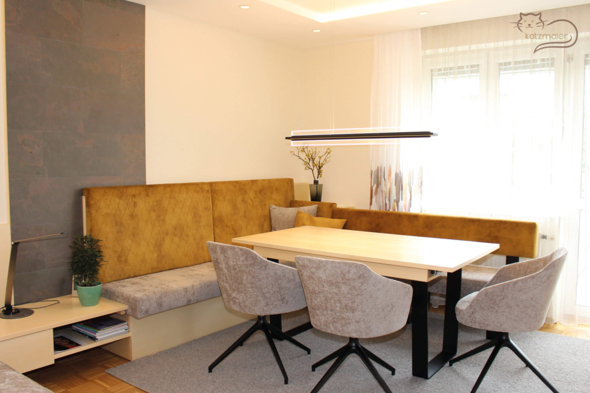 Ein Wohnbereich einem Essbereich. Die Möbel sind in Ahorn furniert kombiniert mit schwarzen, grauen und ockerfarbenen Akzenten. Ein großer Essbereich mit einer Eckbank und drei Stühlen. Die Lehne der Eckbank ist ockerfarben und der Sitz in grau tapeziert. Der Tisch hat eine Platte in Ahorn und ein schwarzes Tischgestell. Über den Tisch hängt eine schwarze gerade Hängeleuchte. Hinter der Eckbank befindet sich eine schmale raumhohe Wandverkleidung in einer Steinoptik.