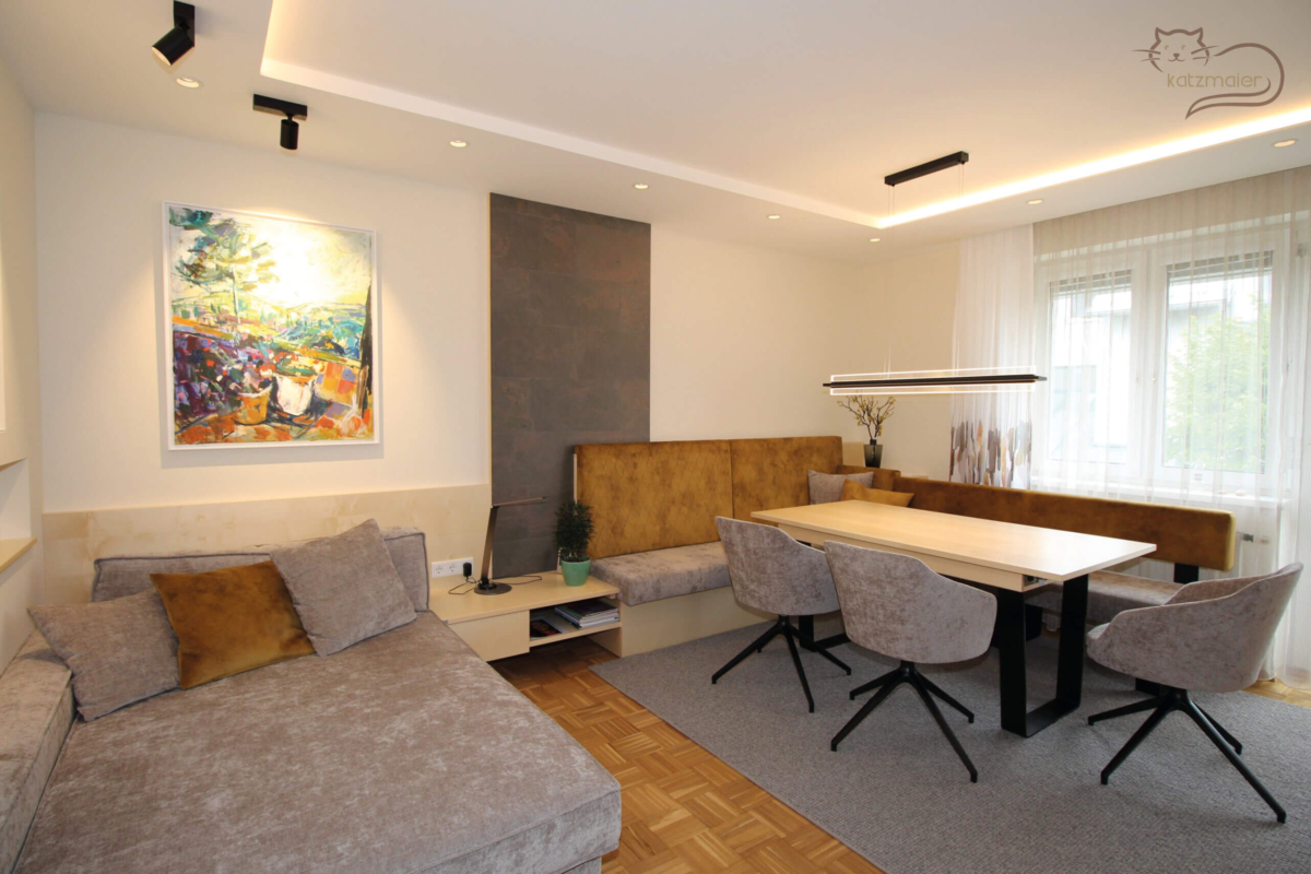 Ein Wohnbereich mit einer Liegecouch und einem Essbereich. Die Möbel sind in Ahorn furniert kombiniert mit schwarzen, grauen und ockerfarbenen Akzenten. Auf der linken Seite im Raum eine graue Liegecouch mit verschiedenen Zierkissen. Dahinter eine Wandverkleidung in Ahorn. Rechts von der Couch eine Ablage mit einer Lade und einer offenen Nische. Anschließend befindet sich ein großer Essbereich mit einer Eckbank und drei Stühlen. Die Lehne der Eckbank ist ockerfarben und der Sitz in grau tapeziert. Der Tisch hat eine Platte in Ahorn und ein schwarzes Tischgestell. Über den Tisch hängt eine schwarze gerade Hängeleuchte. Hinter der Eckbank befindet sich eine schmale raumhohe Wandverkleidung in einer Steinoptik.