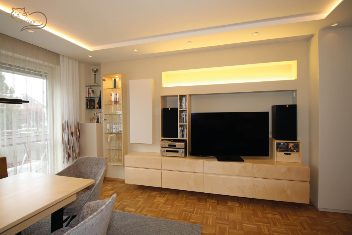 Ein Wohnzimmerverbau in Ahorn kombiniert mit Möbelteilen in Creme. Auf der linken Seite vom Foto ein großes Fenster mit einem weißen Store und einem bunten Vorhang. Entlang der Mauer ein halbhoher Schrank in Creme und darüber zwei Ablageborde. Rechts davon eine aufgehängte Glasvitrine mit innen einer indirekten Beleuchtung. Daneben ein aufgehängter TV-Unterschrank in Ahorn mit Laden. Darüber auf der linken Seite ein aufgehängter Schrank in Creme. Über dem TV-Unterschrank ist eine große Mauernische und darüber eine weitere schmale Mauernische mit einer indirekten Beleuchtung.